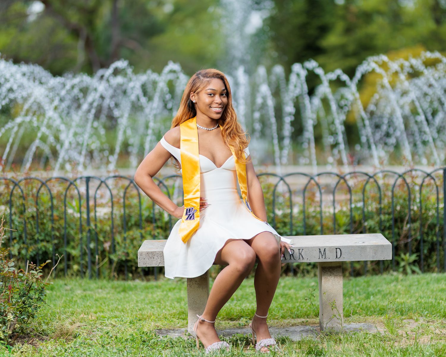 Young woman in white dress and gold graduation sash sits on a bench in front of a fountain outdoors.