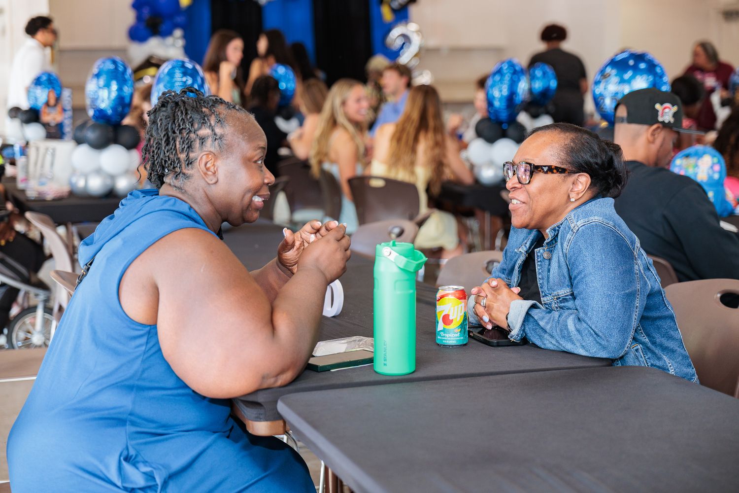 Two women engage in conversation at a table during a blue-themed celebration event.