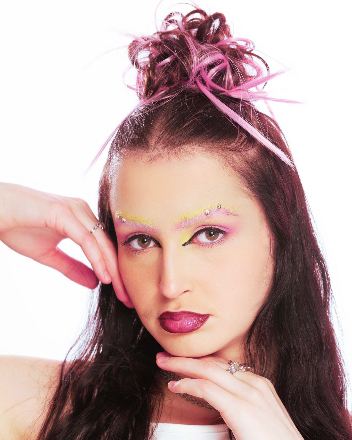 Young woman with dramatic pink and yellow eye makeup, dark lips, and pink-highlighted updo hairstyle against white background.