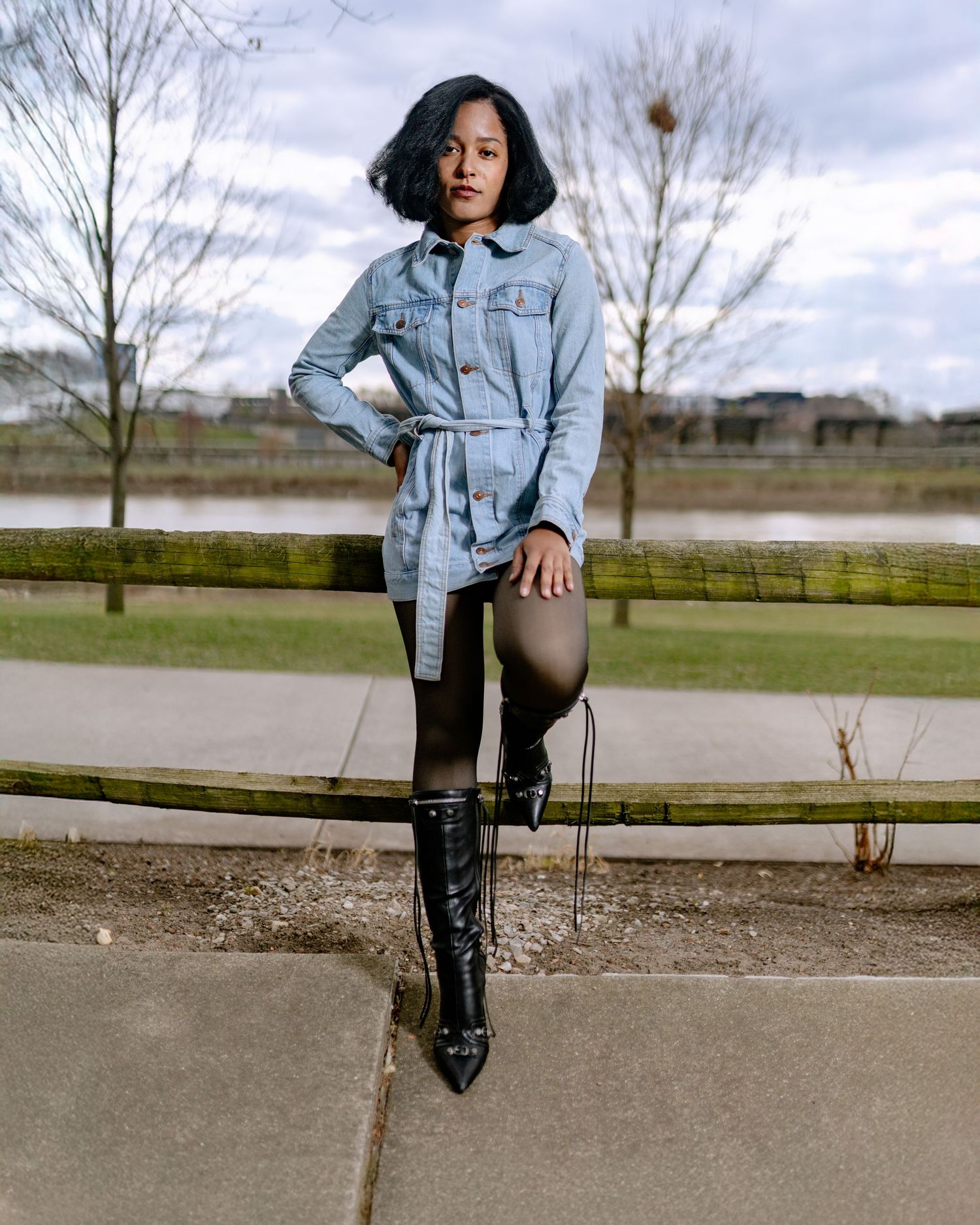 Woman in denim jacket, black leggings, and knee-high boots posing outdoors near a fence.