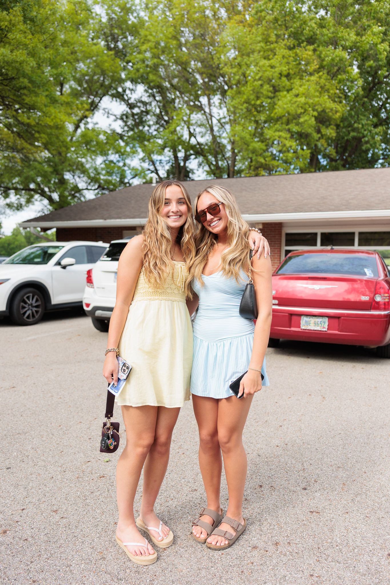 Two young women in pastel summer dresses smiling together in a sunny parking lot.