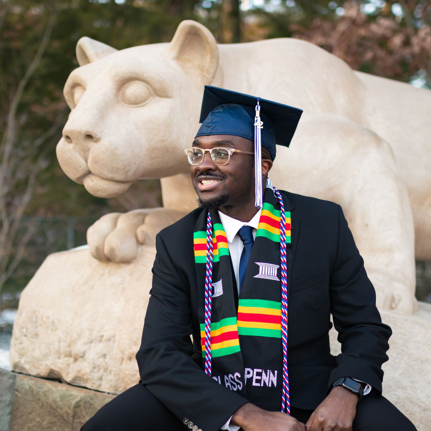 Graduate in cap and gown poses next to iconic Penn State Nittany Lion statue on campus.