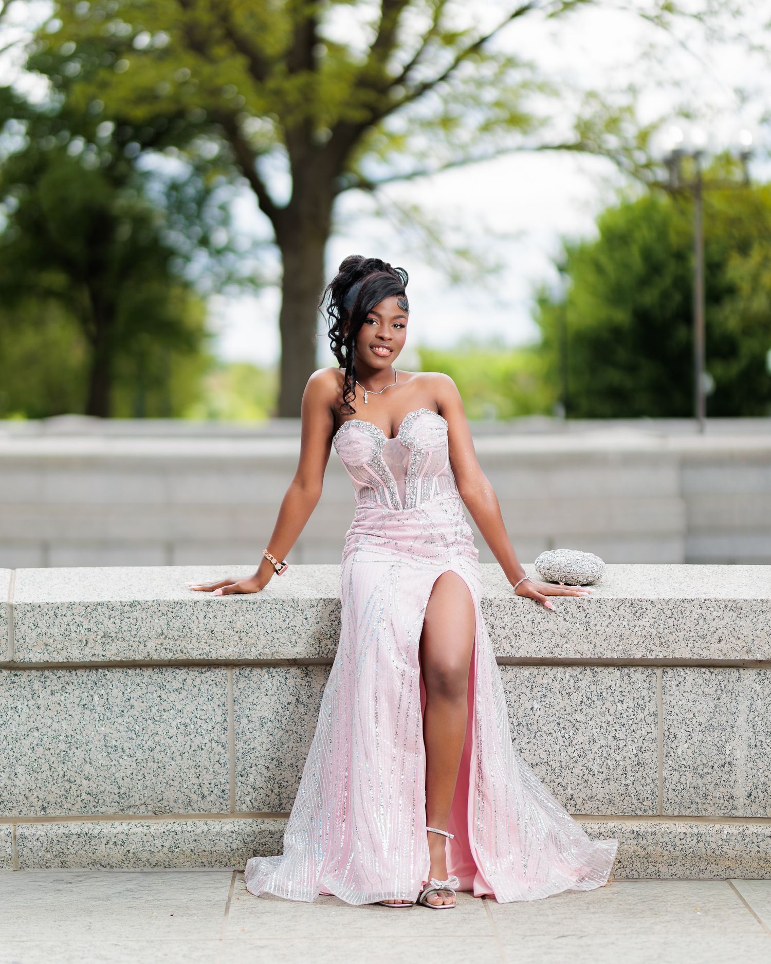 Young woman in pink strapless sequin mermaid prom gown with slit sits on stone steps outdoors.