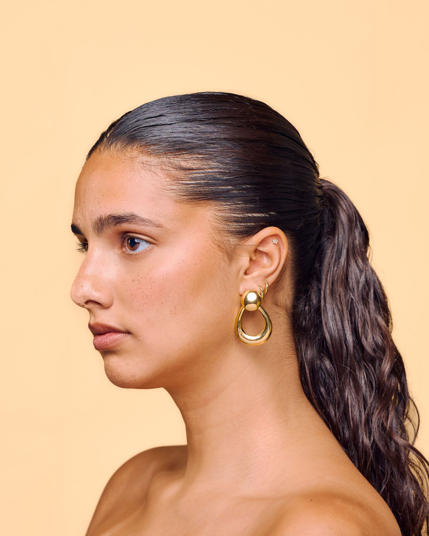Side profile of woman wearing bold gold door-knocker earrings with sleek ponytail on beige background.