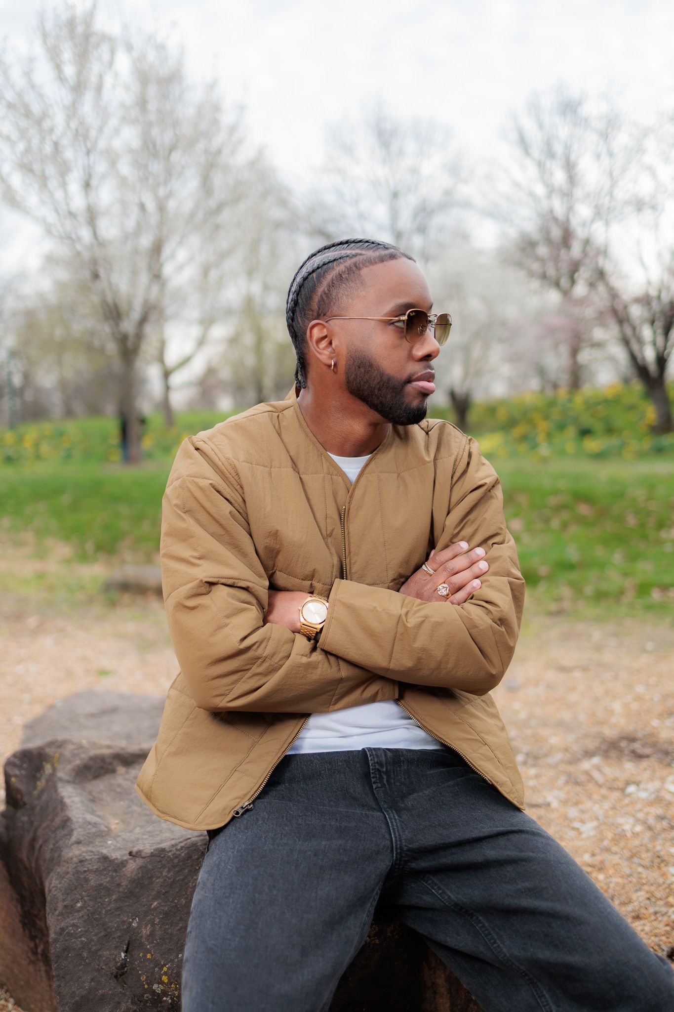 Stylish Black man in tan blazer and sunglasses sits cross-armed on rock in park, looking away thoughtfully.