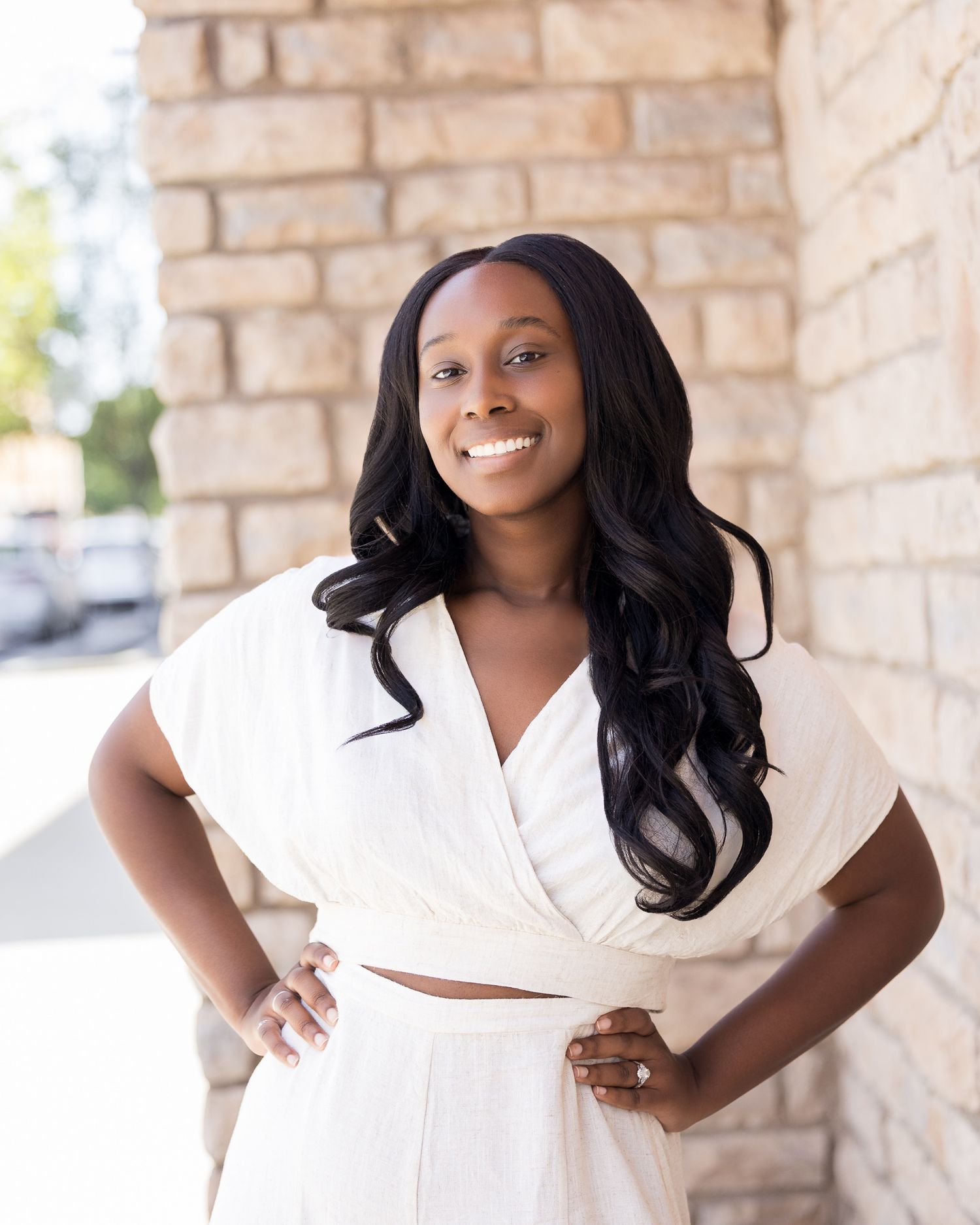 A smiling woman in a white crop top and skirt poses confidently with hands on hips near a stone wall.