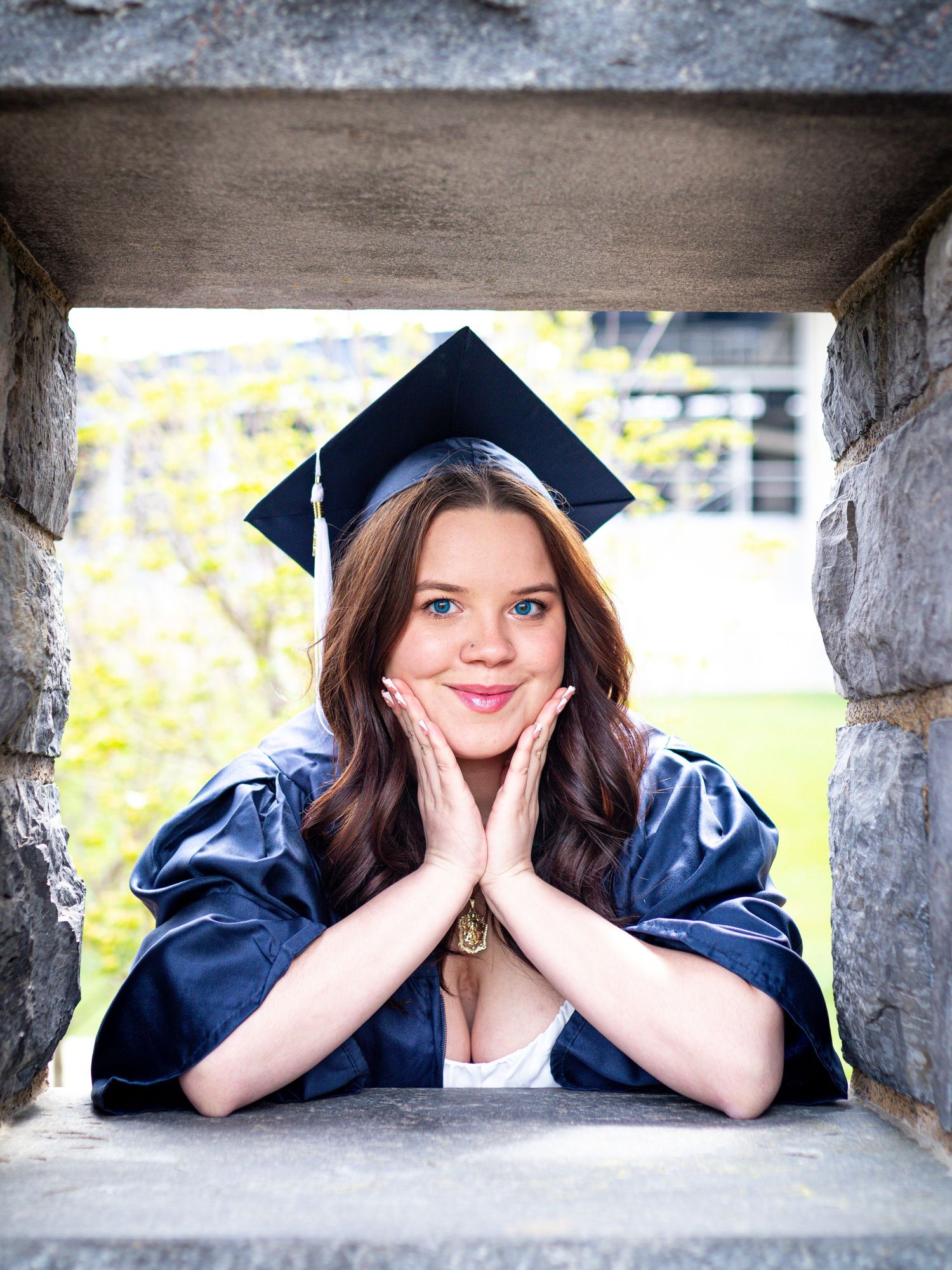 A graduate in navy blue cap and gown rests their elbows in a stone window frame on graduation day.