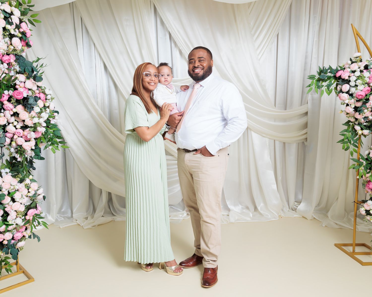 Family portrait with woman in mint dress, man in white shirt, and baby posed before floral arch backdrop.