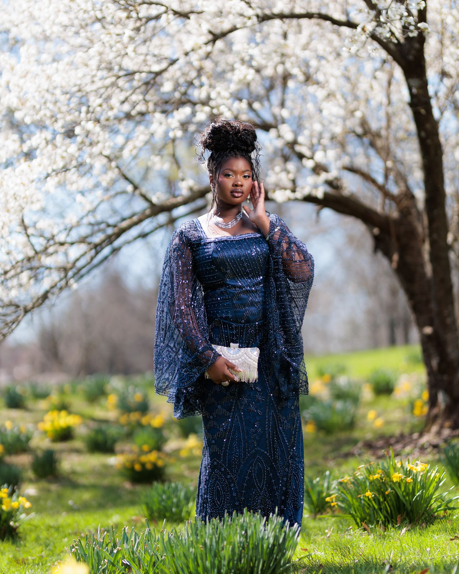 A young woman in a navy sequined gown with sheer sleeves poses outdoors among spring blooms and daffodils.