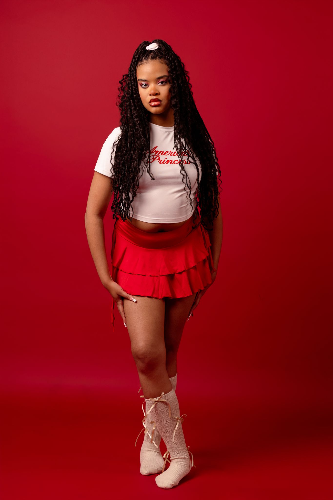 Fashion portrait against red backdrop showing long dark curls, white crop top, red ruffled skirt and lace-up ballet flats.