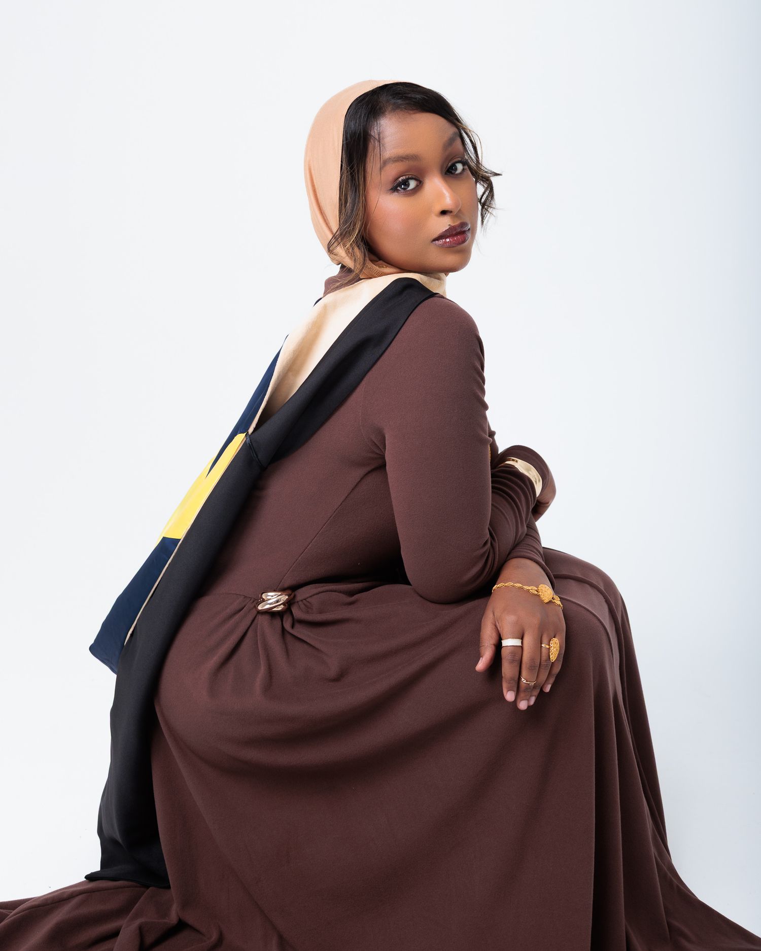 Woman in brown abaya and beige hijab with black and yellow sash poses gracefully against white background.