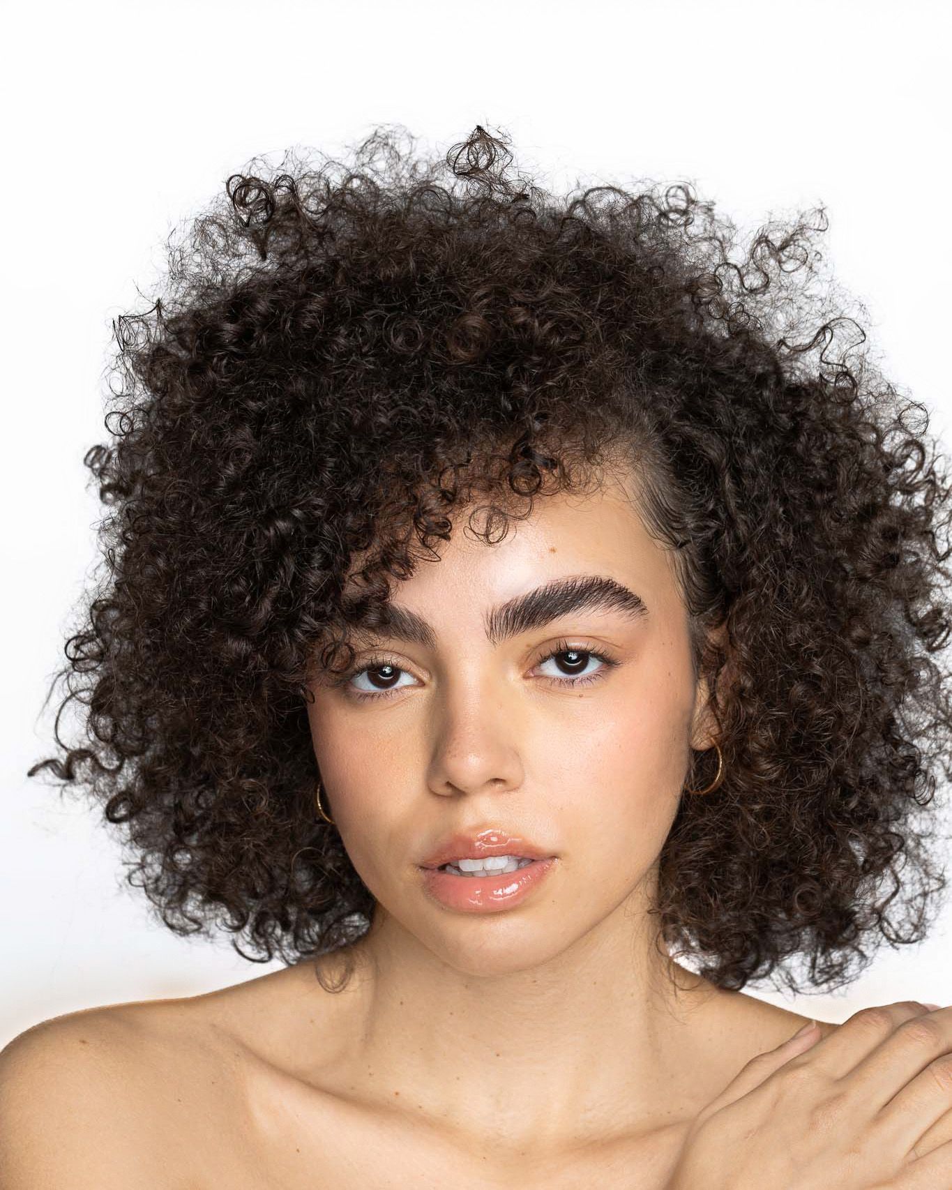 Young woman with voluminous dark curly hair, bold brows, and natural makeup poses against a white background.