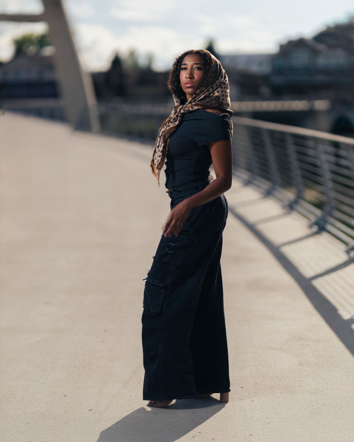 Woman in black maxi dress and leopard print headscarf poses confidently on a modern bridge walkway.