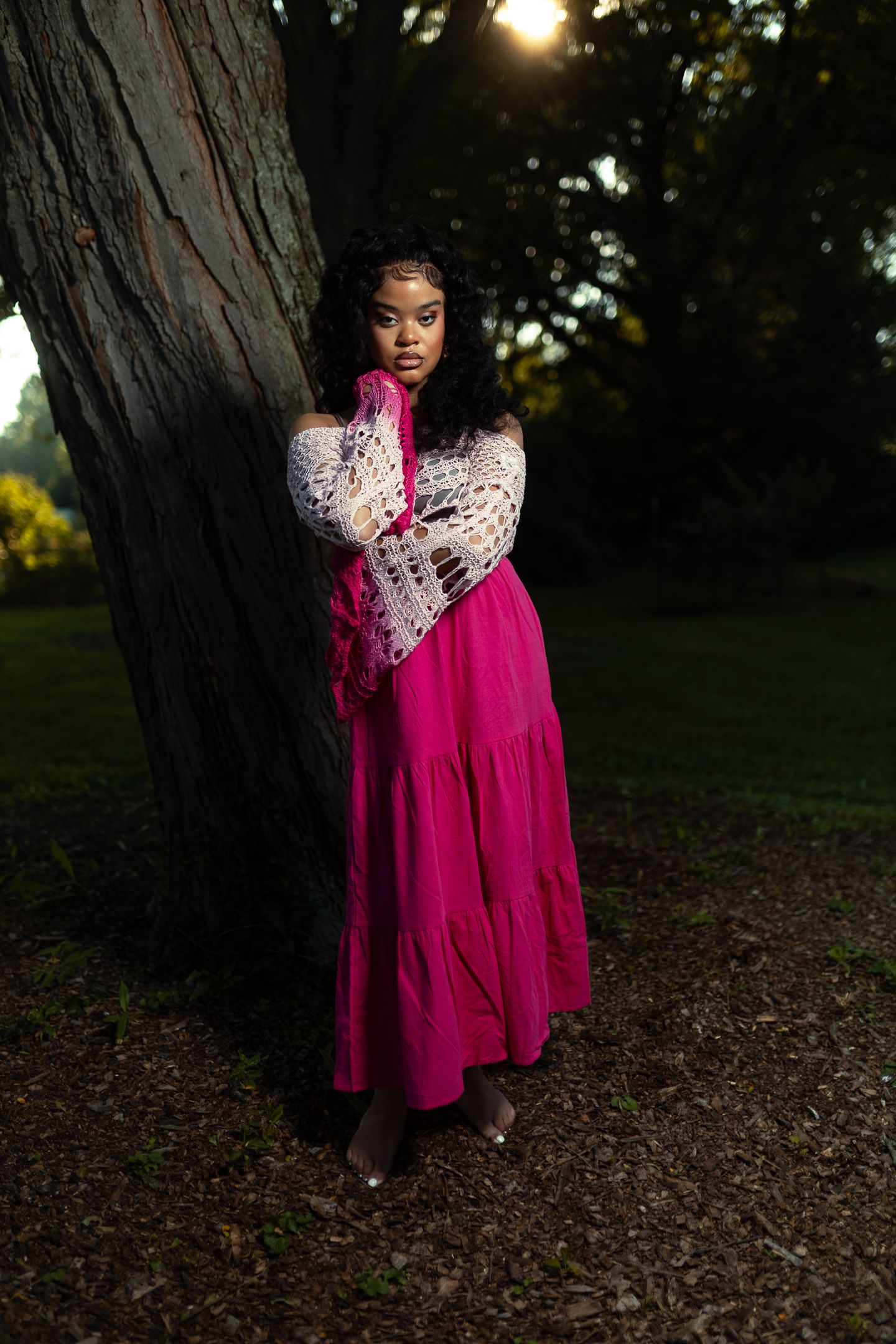 A young Black woman in a pink maxi skirt and white lace top leans against a tree in a sunlit park.
