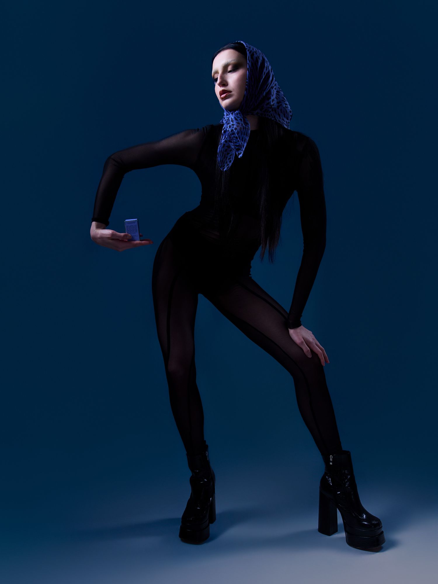Model in black bodysuit and platform boots poses dramatically against blue backdrop, wearing purple headscarf.