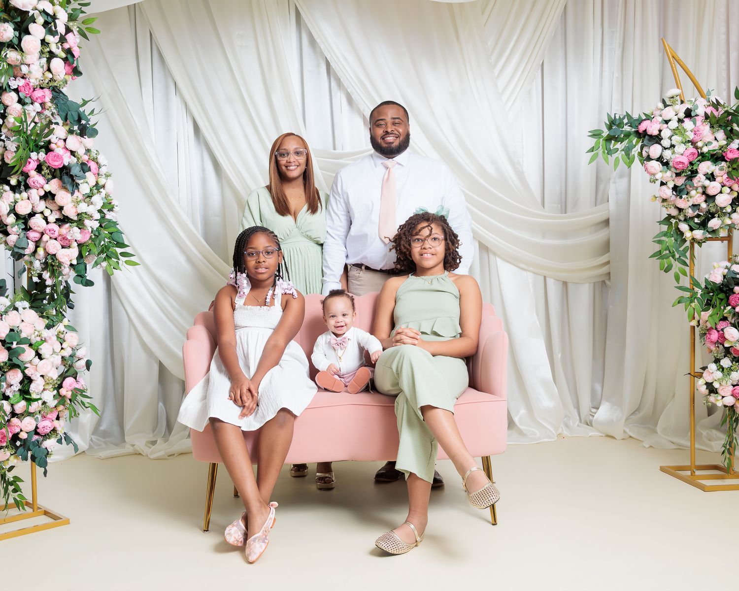 Family of five posing elegantly in a floral studio setting with pink roses and white draping backdrop.
