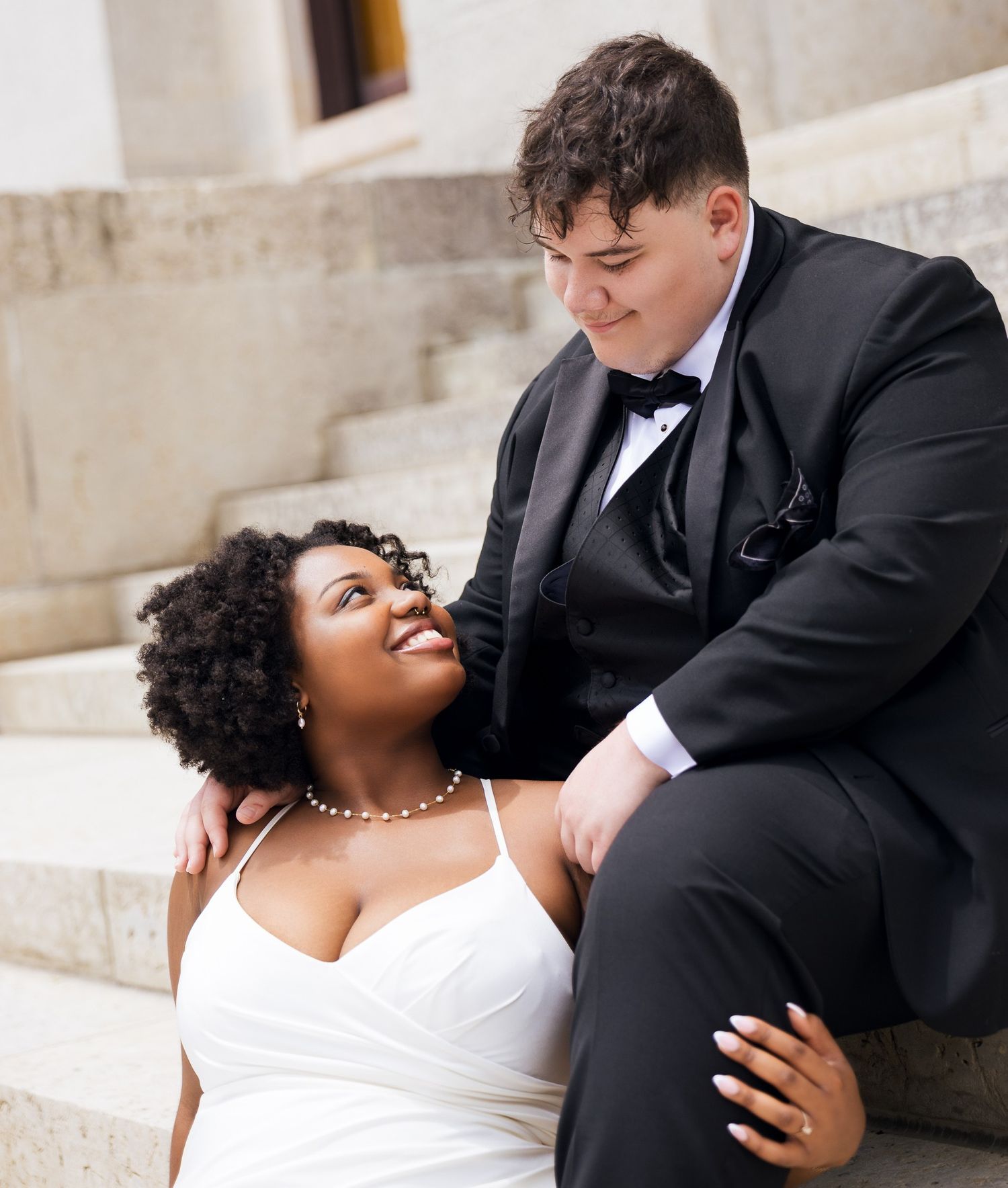 Interracial couple in wedding attire sitting on stone steps, sharing a tender romantic moment.