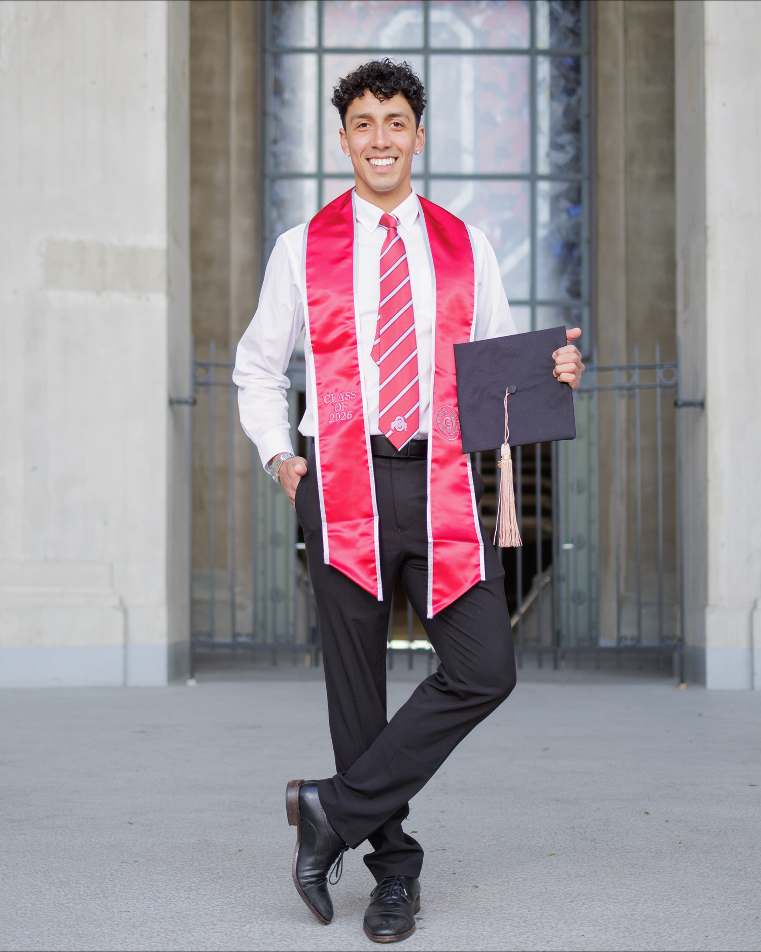 Smiling graduate in red sash and striped tie holds cap outside university building.