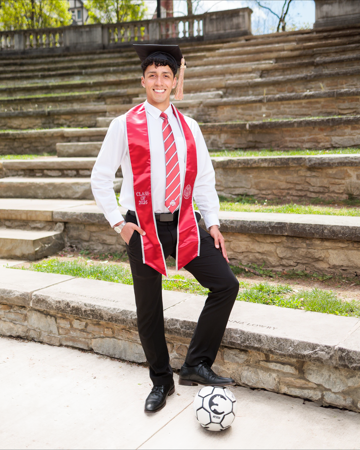 Smiling graduate in cap, red stole, and tie poses with soccer ball on stone steps outdoors.