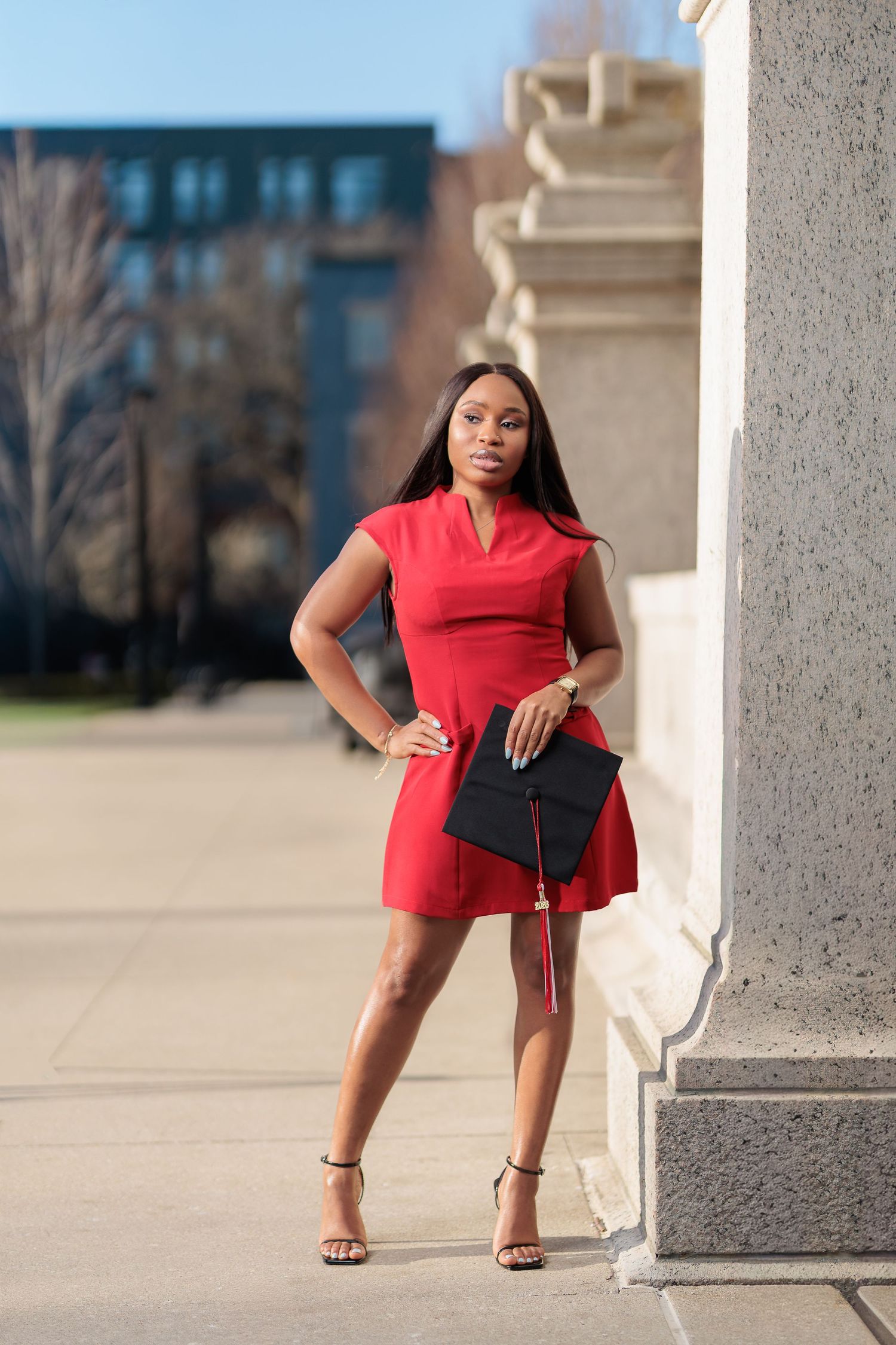 Young Black woman in red dress holding graduation cap poses confidently by stone column outdoors.