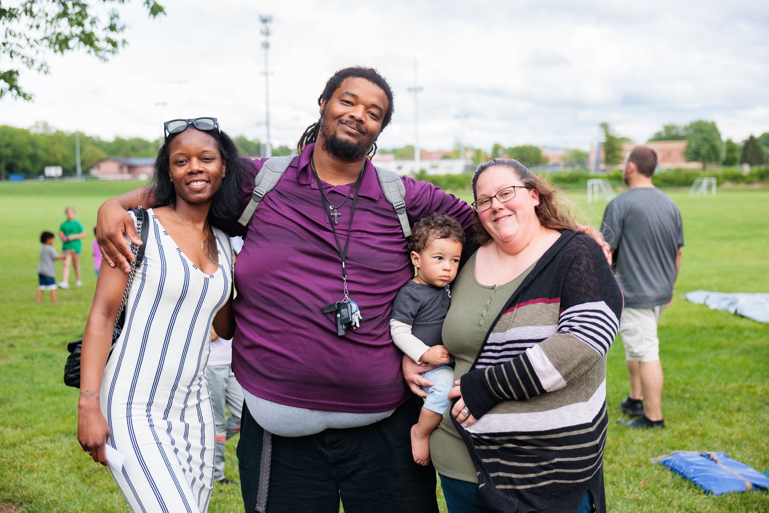 Family of four smiles together outdoors at a community event on a green field.
