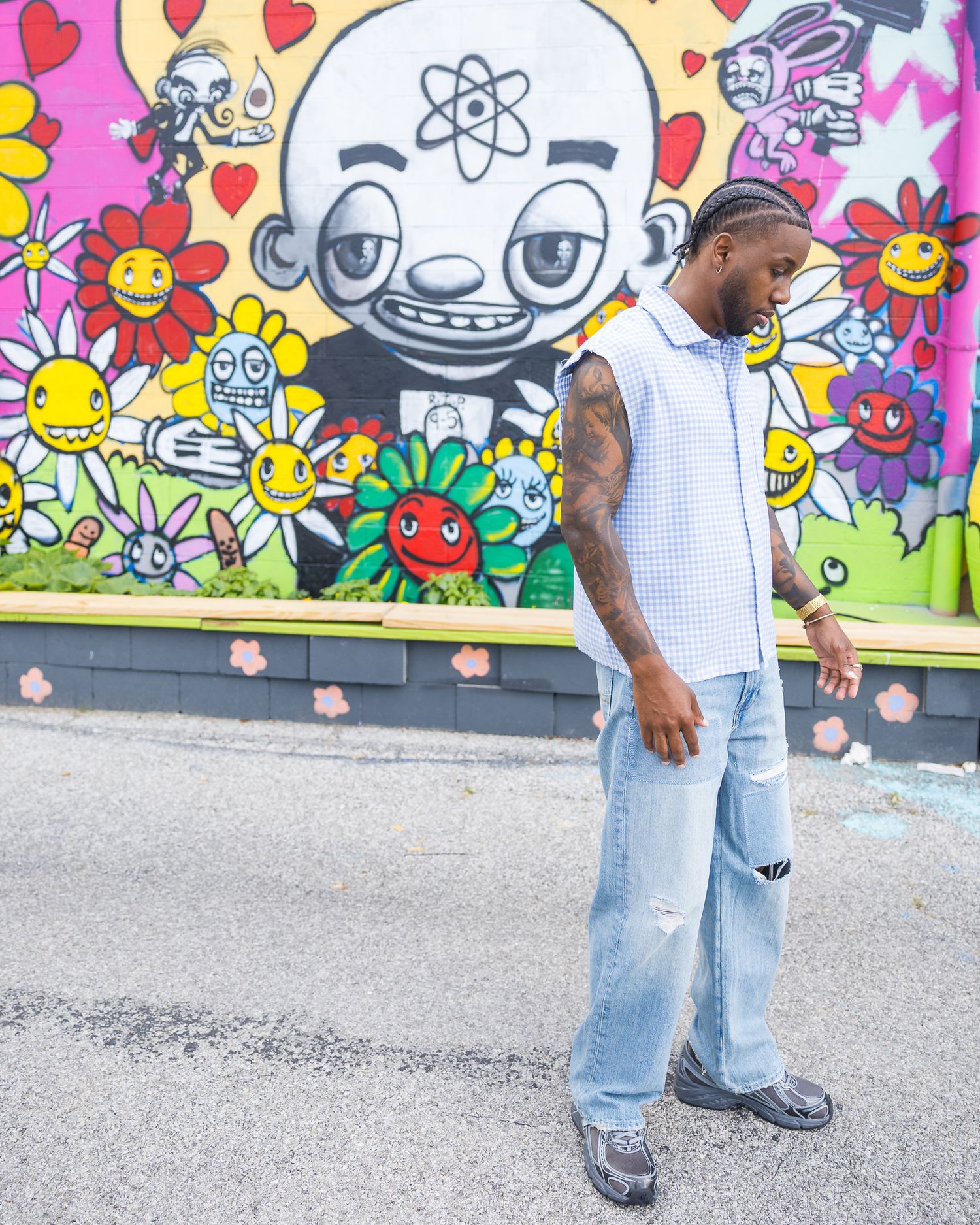 Man in sleeveless shirt and baggy jeans posing in front of colorful street art mural with cartoon characters.