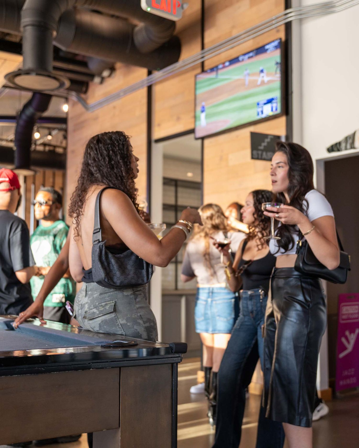 People gather at a casual indoor venue with exposed wood walls and industrial decor while a sports game plays on TV.