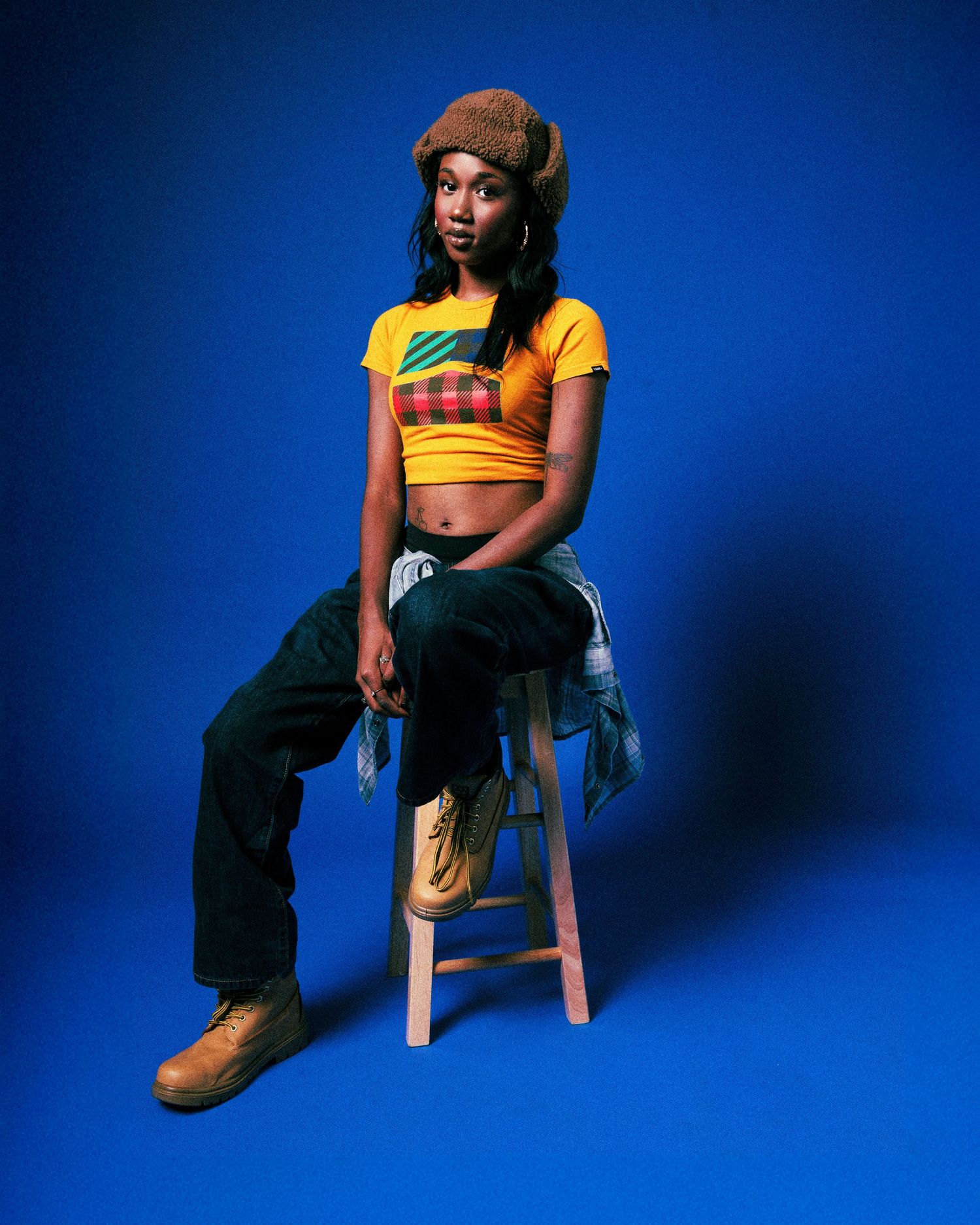 Person in a yellow crop top and brown pants sits on a wooden stool against a vibrant blue studio backdrop.