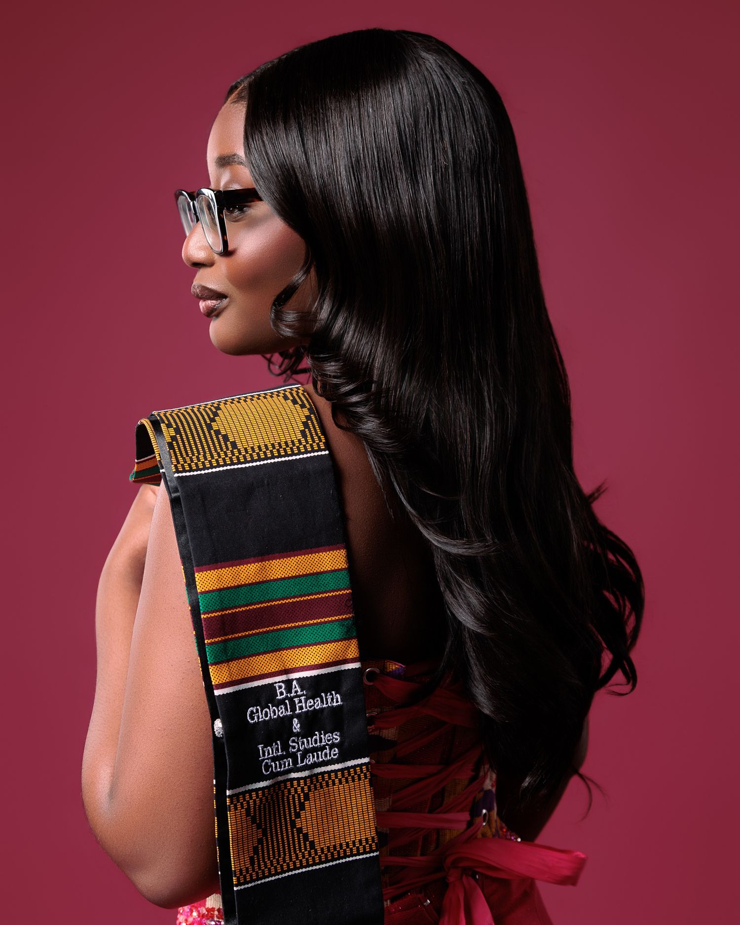 Graduate in kente stole poses with long black hair, glasses, and B.A. Global Health degree sash against pink background.