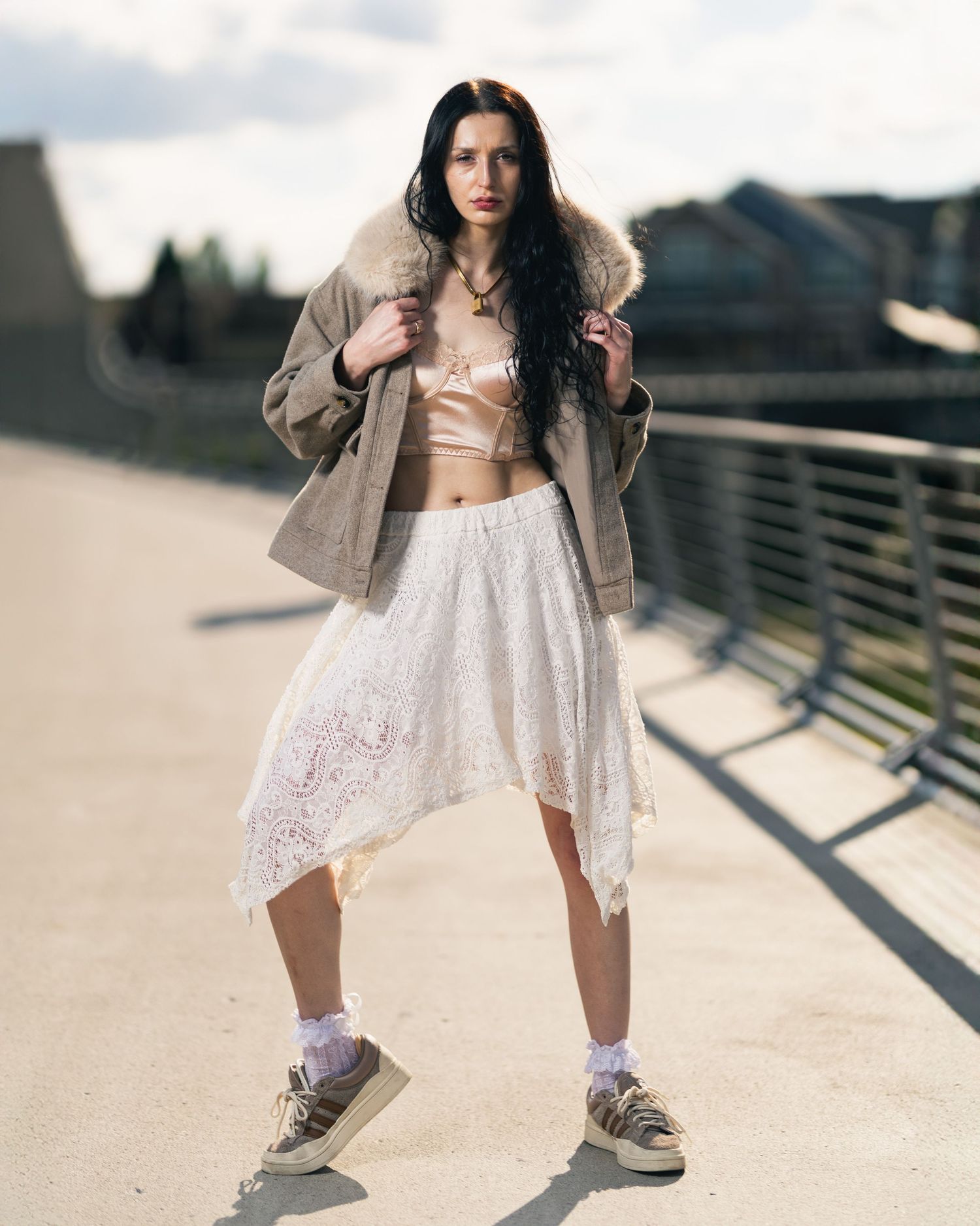 Young woman in lace skirt, crop top, and oversized jacket poses confidently on a sunny urban bridge.