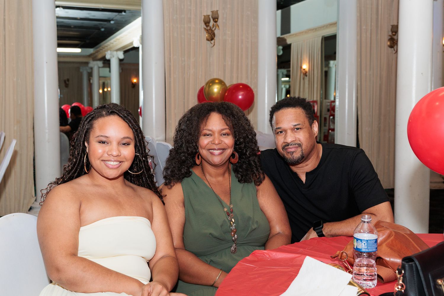 Three people smiling together at a festive event with red balloons in the background.