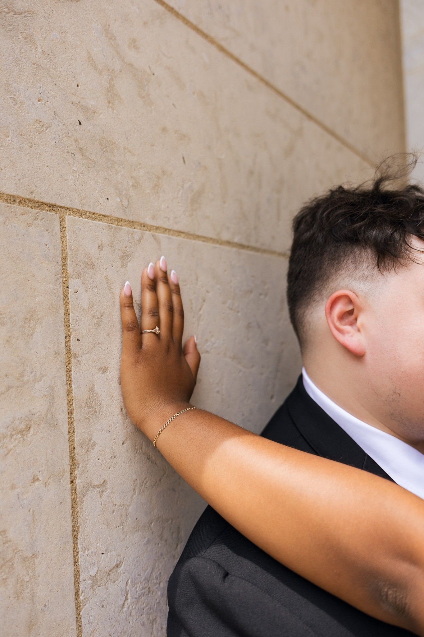 Bride's hand with engagement ring pressed against marble wall beside groom in black suit at wedding venue.
