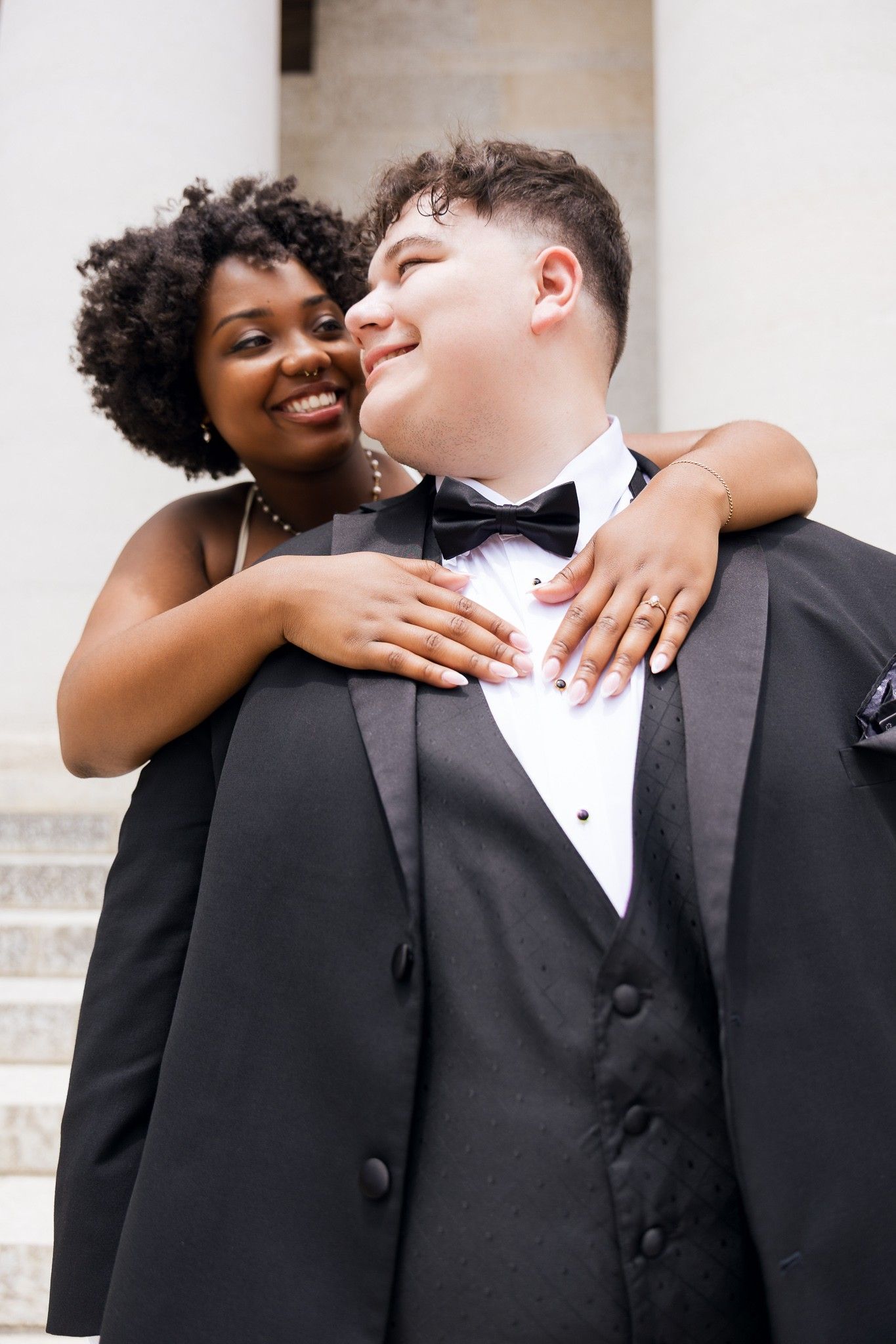 A young interracial couple in formal attire, woman in dress embracing man in tuxedo, smiling outdoors.