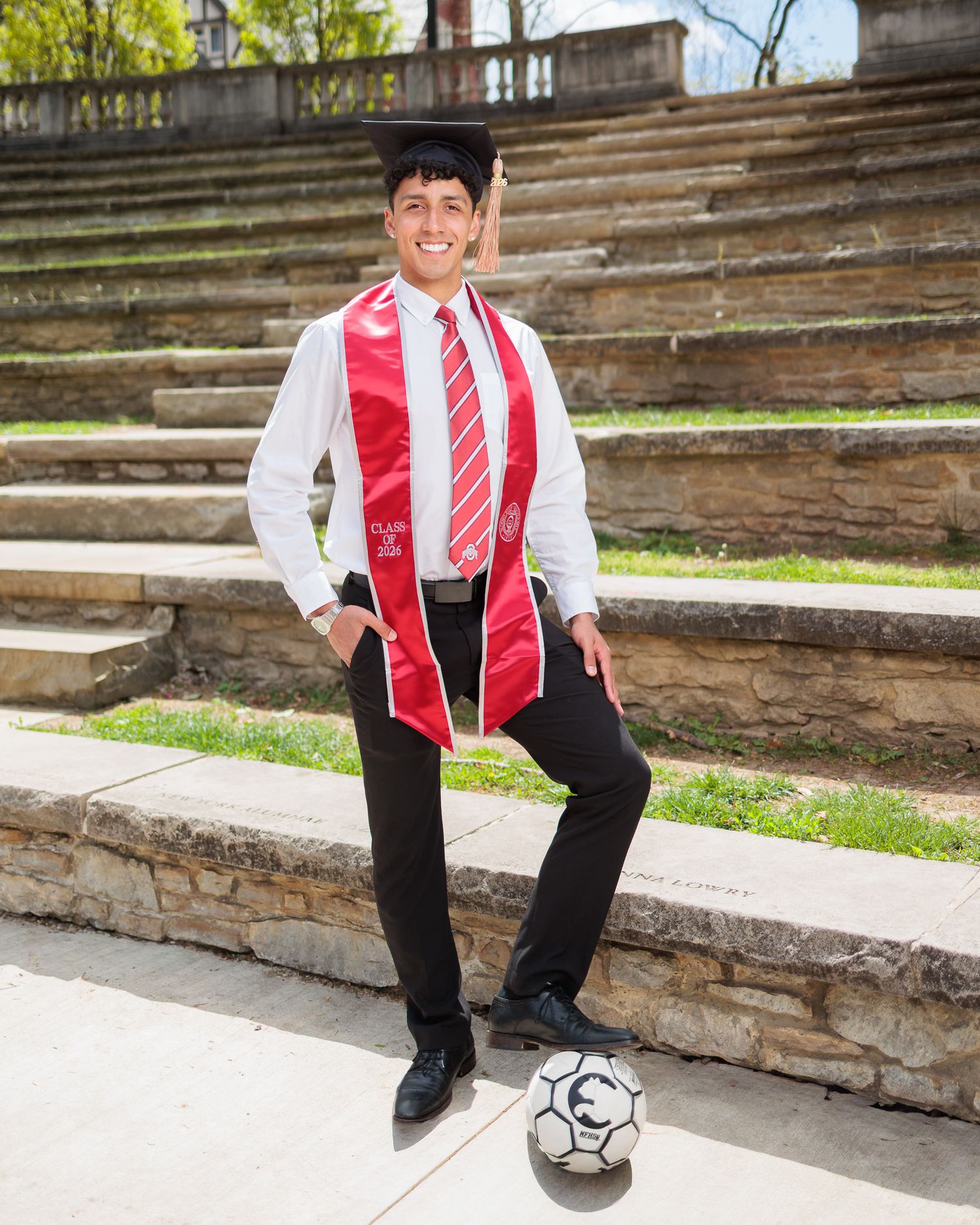 Graduate in cap and red stole poses with soccer ball on stone steps outdoors.