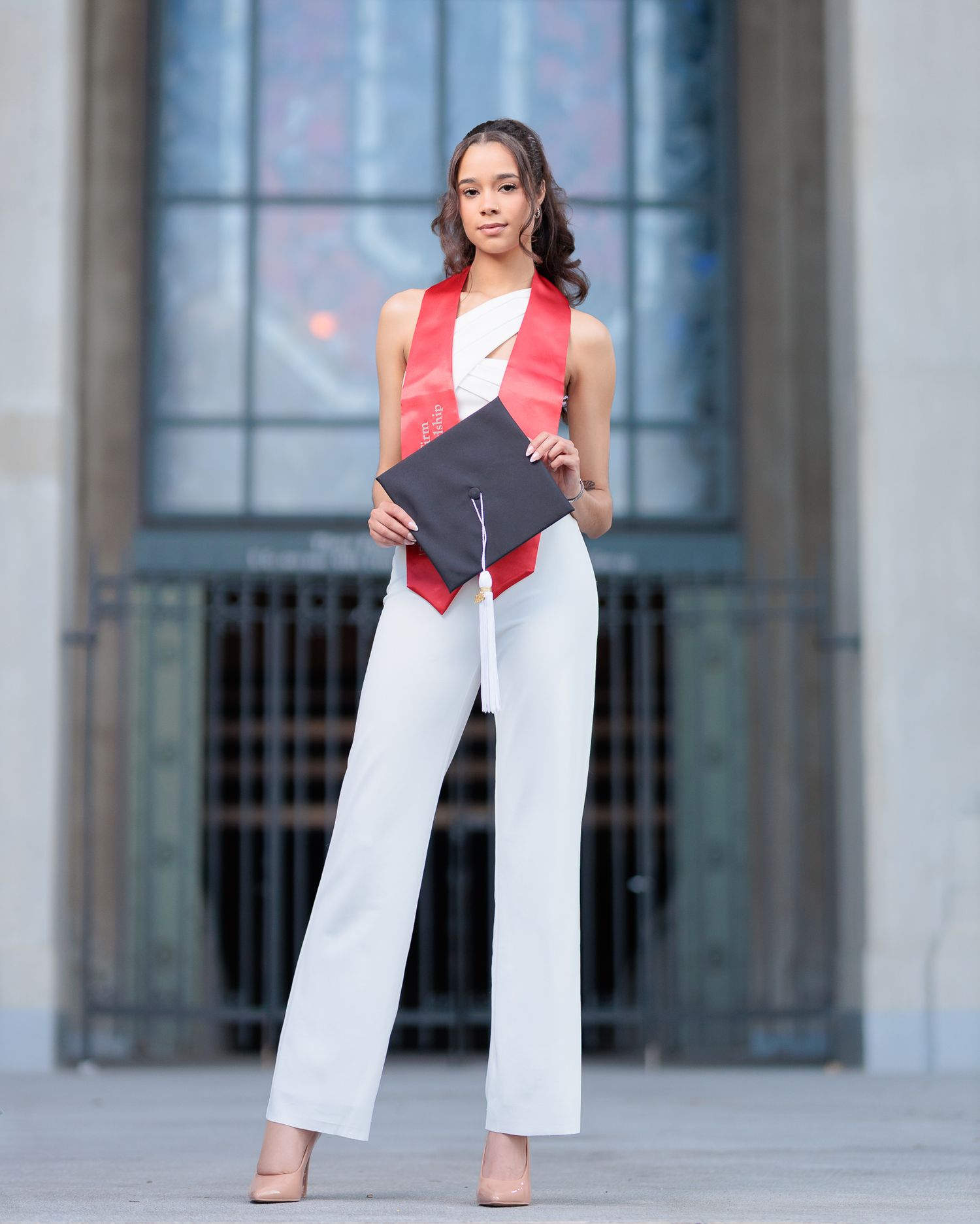 Young woman in white pants and red graduation stole holds cap, posing proudly in front of a grand building.