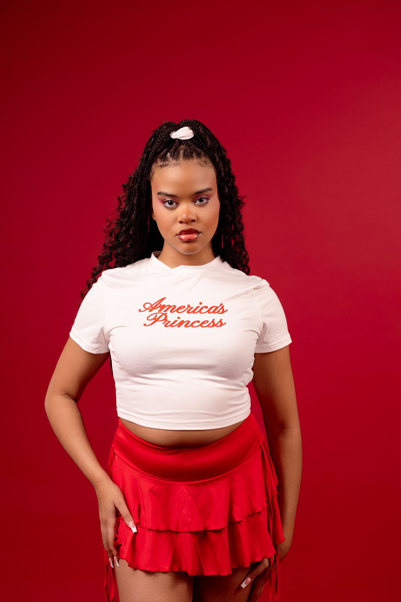 A figure in a white cropped t-shirt reading 'American Princess' and red pleated mini skirt poses against a red backdrop.