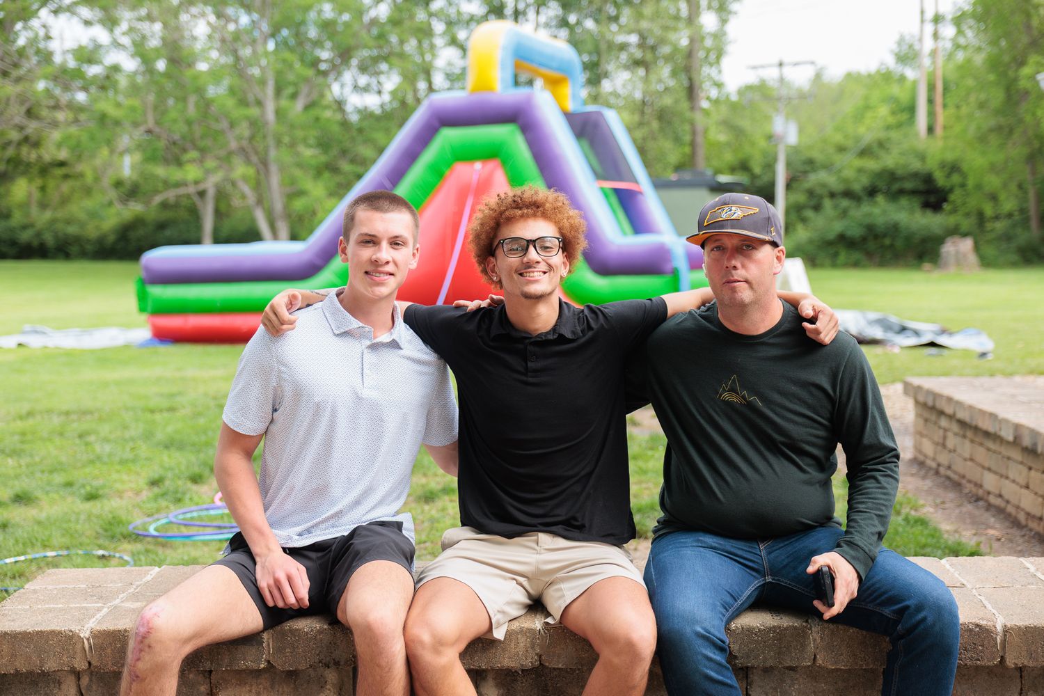 Three smiling men pose together outdoors near a colorful inflatable bounce house at a summer party.