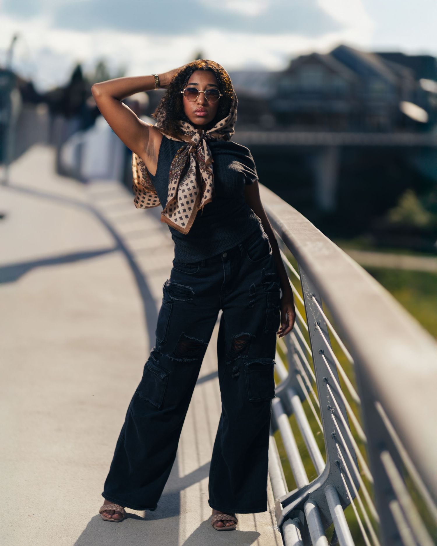 Stylish woman in black wide-leg pants and leopard print head scarf posing on a bridge railing.