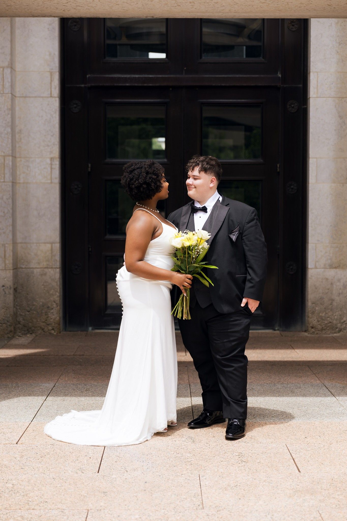Interracial newlyweds pose before ornate doors, bride in white gown holding yellow flower bouquet.