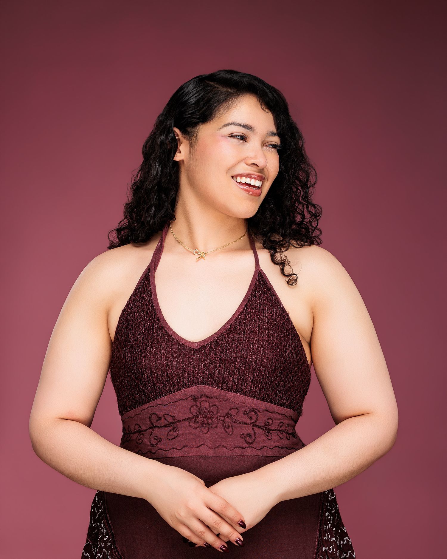 Smiling woman in a burgundy halter dress with curly black hair poses against a mauve background.