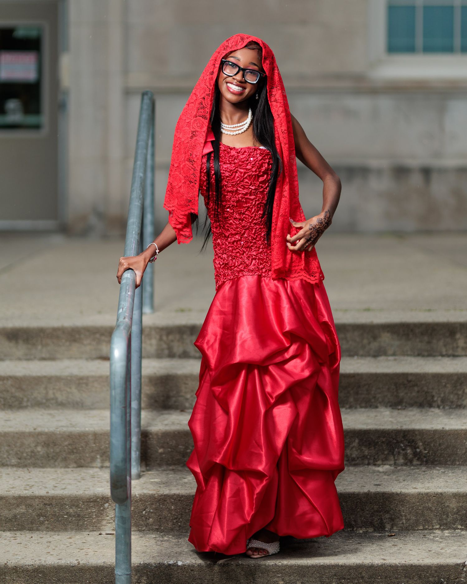 A woman poses gracefully in a stunning red mermaid gown with matching lace veil on steps.