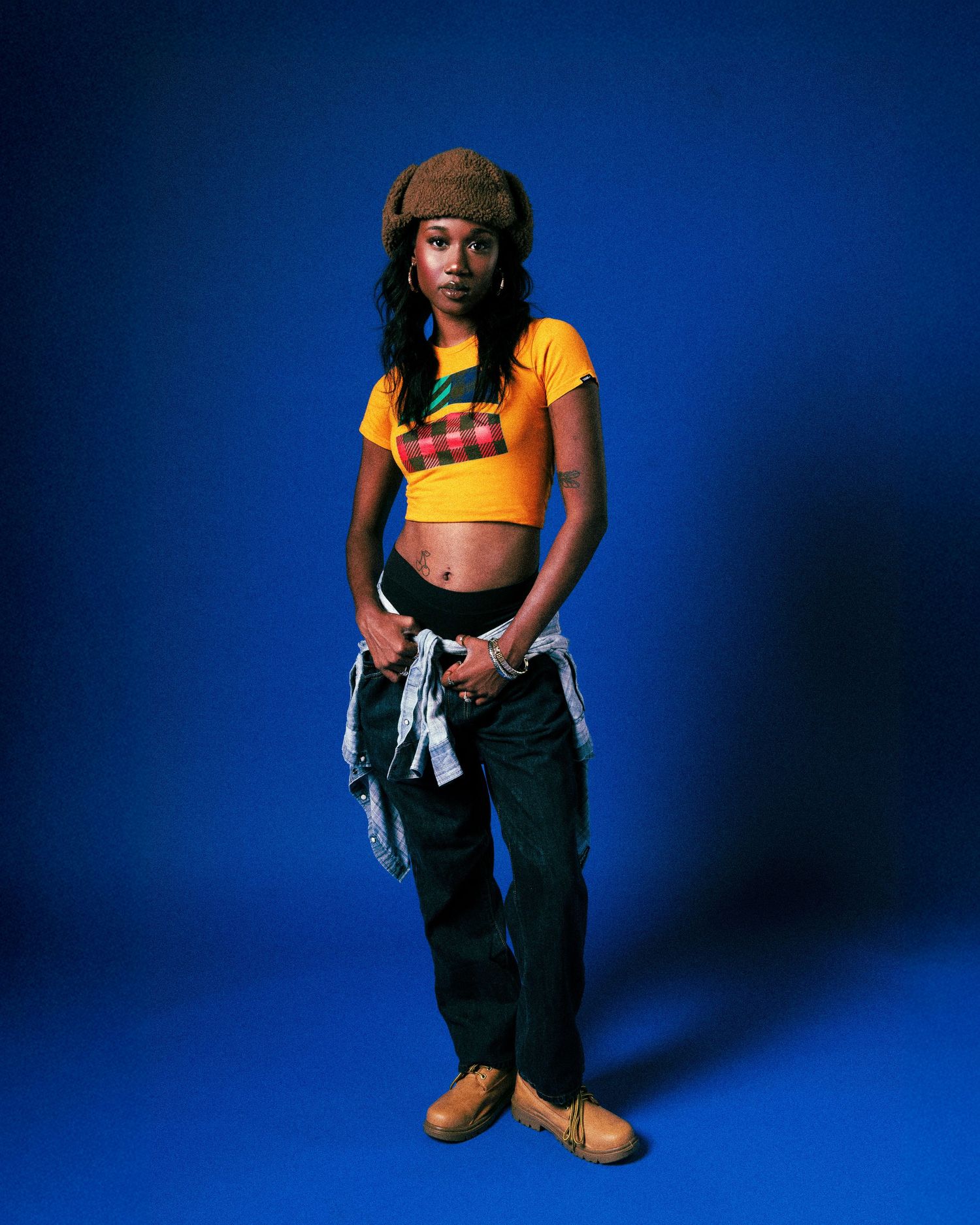 A figure in a yellow crop top and dark pants poses against a deep blue studio backdrop.