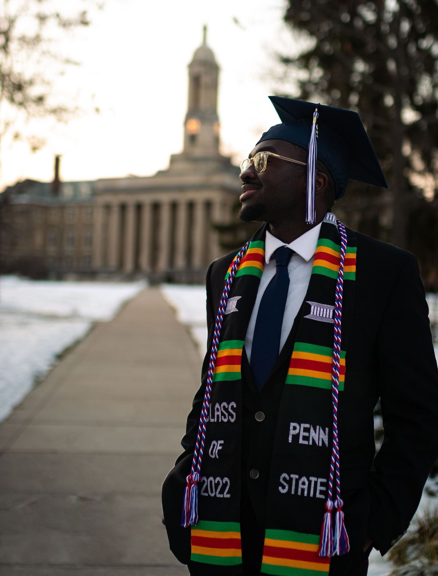 Graduate in cap and kente stole standing in front of Penn State campus building at sunset.