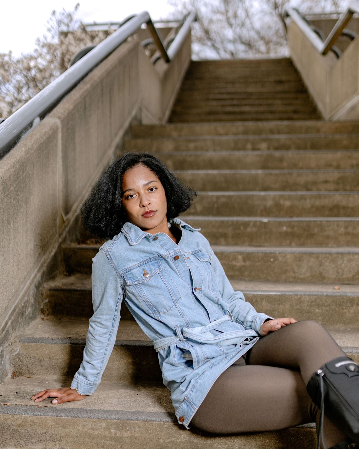 Young woman in denim jacket sitting on concrete stairs outdoors, with curly hair and brown tights.