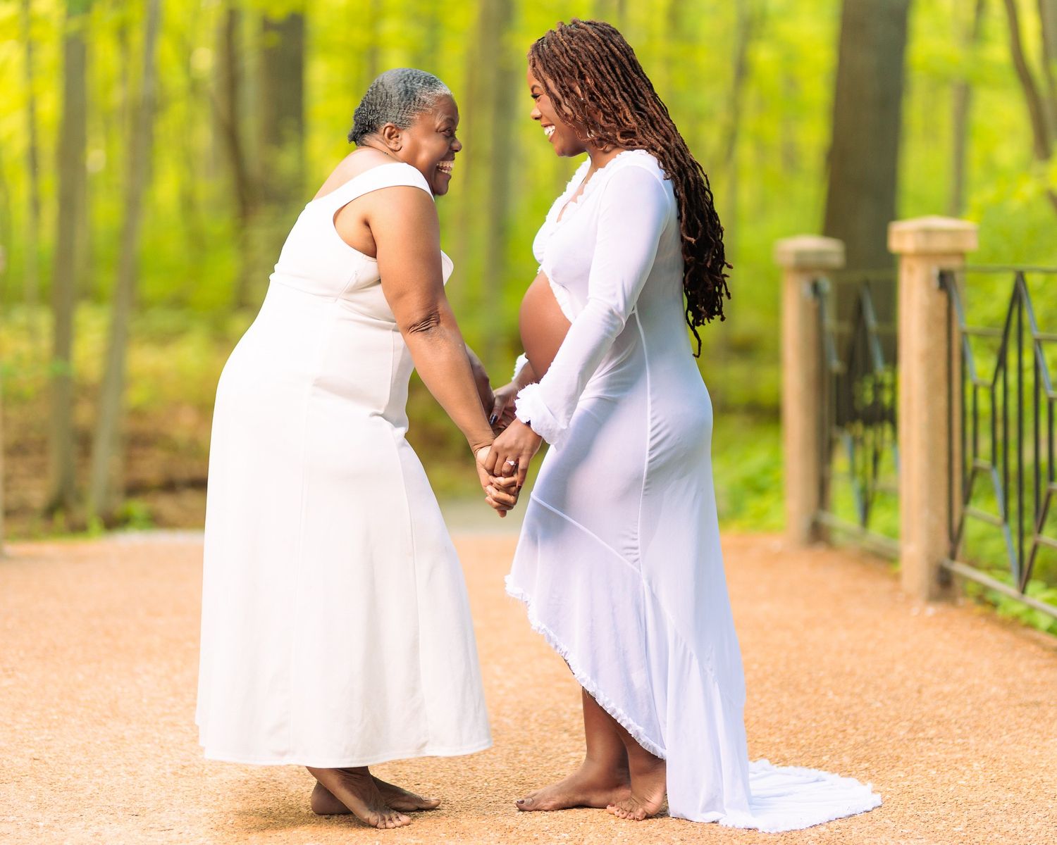Two women in white dresses hold hands outdoors, one pregnant, sharing a tender moment on a sunlit path.