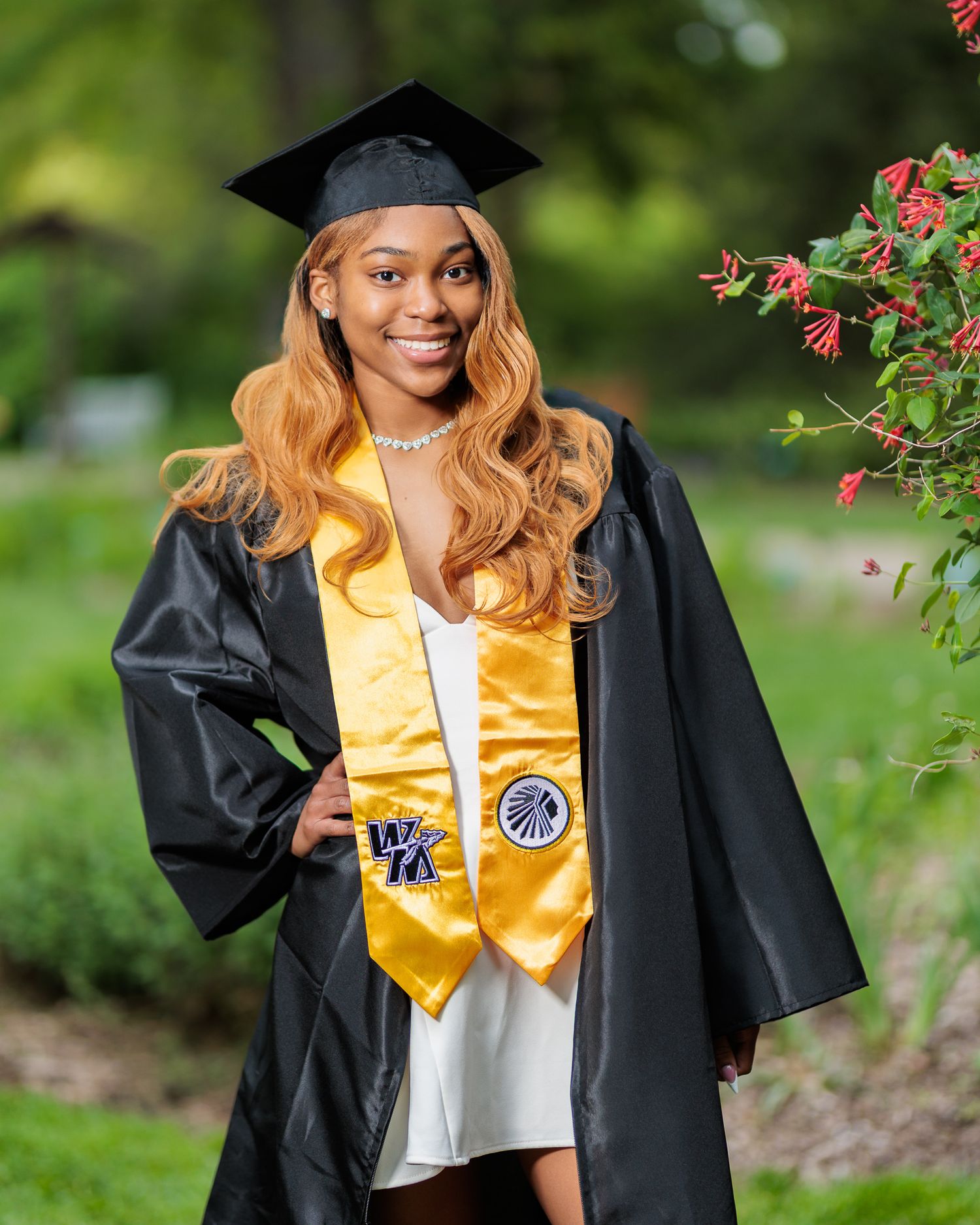 Young woman in black graduation cap and gown with gold stole smiles outdoors near red flowers.