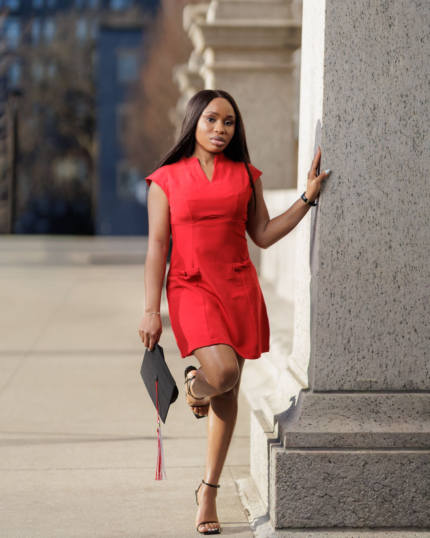 Young woman in red dress holding graduation cap poses confidently by a column outdoors.