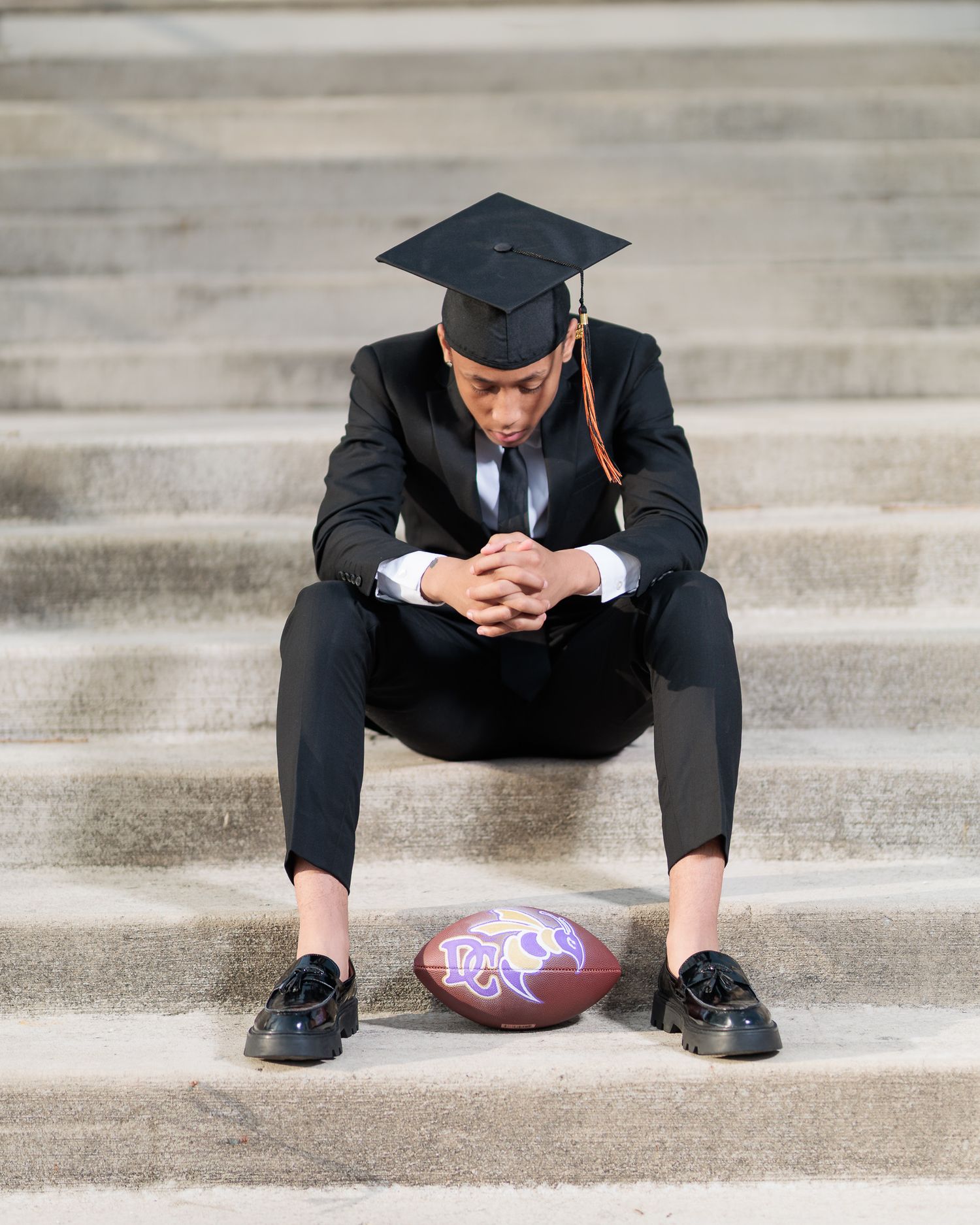Graduate in black suit and cap sits on steps, gazing down at a purple football beside his feet.
