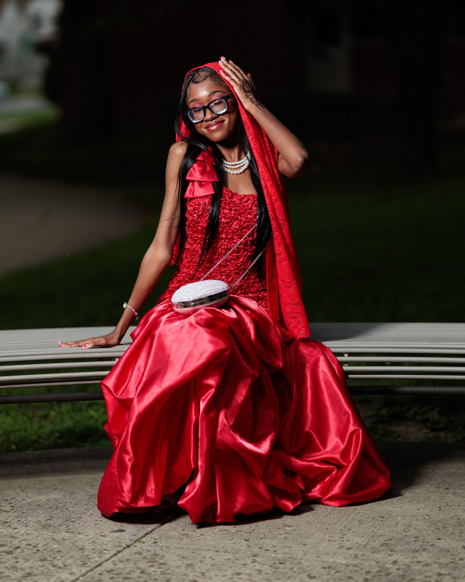 Young woman in elegant red ballgown with cape and glasses poses gracefully on bench at night.