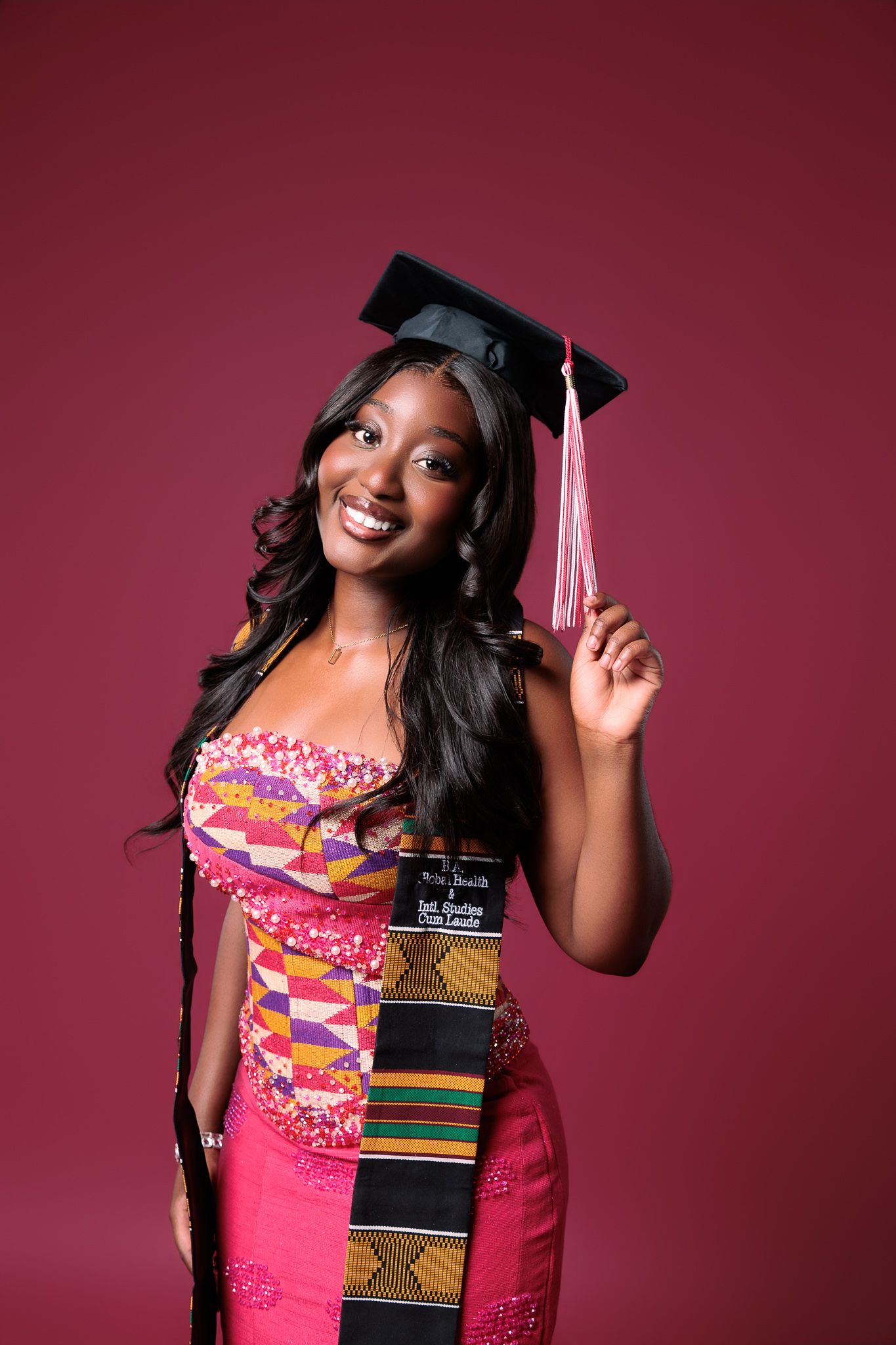 Smiling graduate in pink kente stole and mortarboard cap holding tassel against burgundy background.